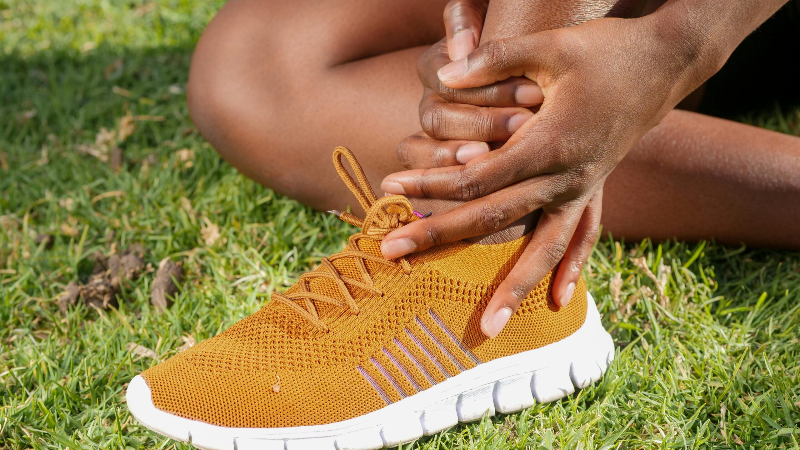 Close-up of athlete holding ankle with running shoe on grass, indicating pain or injury.